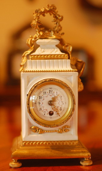 A miniature French timepiece with ormulu decorations on a white Bisque case. A convex enamelled dial showing garlands of flowers around Arabic numerals . The case design is asymmetric and stands on a gilt base . Circa 1890-1900 .